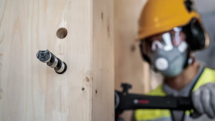 Een persoon met veiligheidsuitrusting boort een gat in een houten balk.