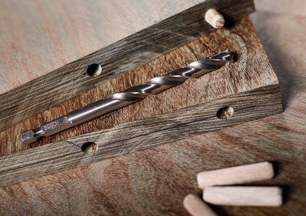 Boortje ligt op houten planken met geboorde gaten en losse deuvels ernaast.