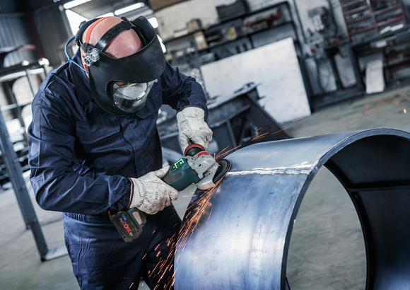 Een persoon die veiligheidsuitrusting draagt, slijpt tegen de rand van een grote metalen pijp, waardoor vonken ontstaan.