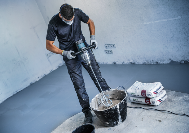 Een persoon met veiligheidsuitrusting mengt cement in een emmer met behulp van een elektrische mixer.