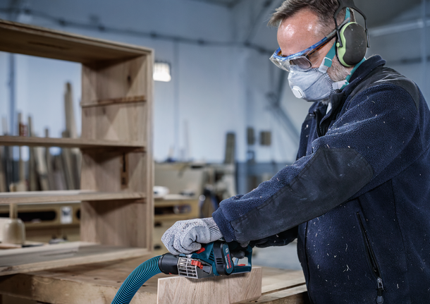 Een persoon met veiligheidsuitrusting schuurt een houten plank in een werkplaats.