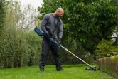 Een persoon met veiligheidsuitrusting maait het gras langs de tuinrand met een snoerloos gereedschap.