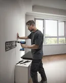Een persoon die veiligheidsuitrusting draagt, monteert een metalen beugel aan een muur met behulp van een accuboormachine.