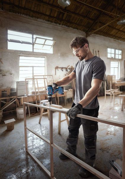 Een persoon met veiligheidsuitrusting boort in een houten frame in een werkplaats.