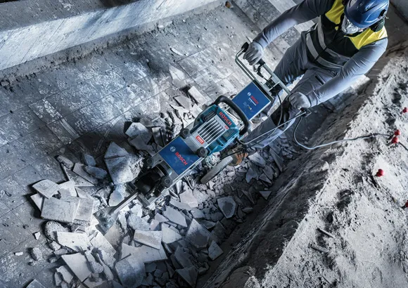 Een persoon met veiligheidsuitrusting breekt een betonnen vloer af met behulp van een zwaar sloopgereedschap.