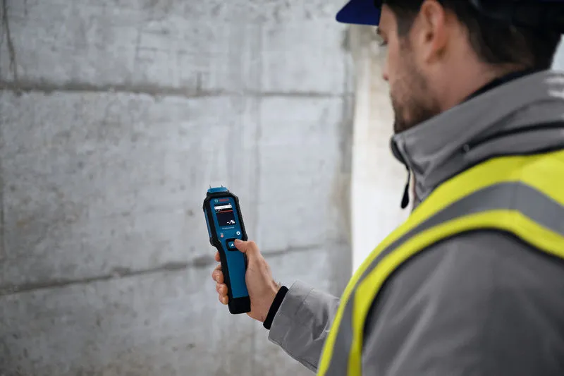 Een persoon die veiligheidsuitrusting draagt, controleert met een materiaalvochtigheidsmeter een betonnen muur.