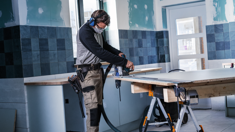 Een persoon die veiligheidsuitrusting draagt, gebruikt een elektrisch gereedschap om een plank te zagen in een betegelde kamer.