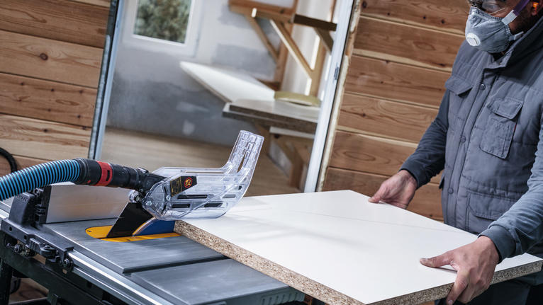 Een persoon die veiligheidsuitrusting draagt, bedient een tafelzaag om een houten plank te zagen.