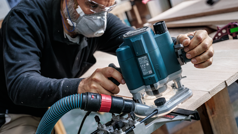 Een persoon die veiligheidsmateriaal draagt, gebruikt een bovenfrees om de rand van een houten plank vorm te geven.