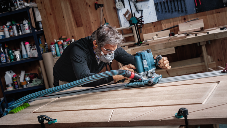 Een persoon die veiligheidsuitrusting draagt, gebruikt een elektrisch gereedschap om groeven in een houten deur te frezen.