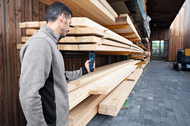 Een persoon meet de vochtigheidsgraad van gestapeld hout in een houtopslagruimte met een materiaalmeter.