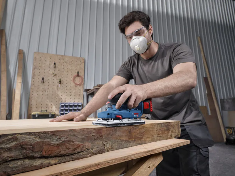 Een persoon met veiligheidsuitrusting schuurt een houten plank met een snoerloze excentrische schuurmachine.