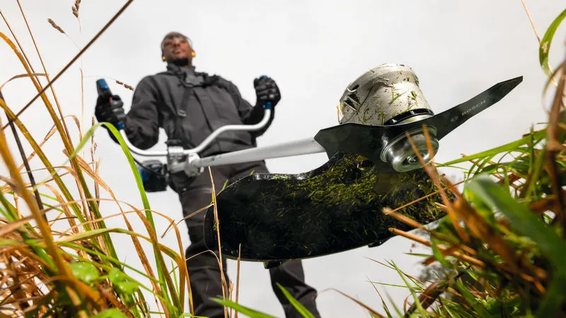 Een persoon met veiligheidsuitrusting maait hoog gras met een bosmaaier.