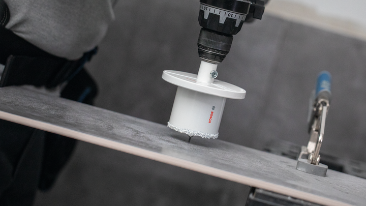 Een persoon met veiligheidsuitrusting boort een gat in een keramische tegel.