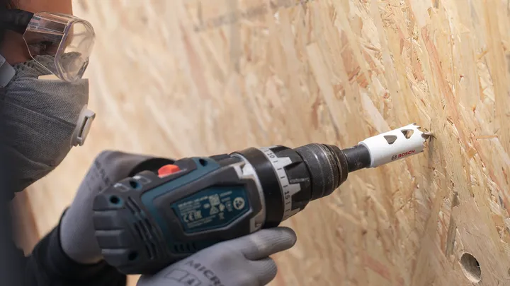 Een persoon met veiligheidsuitrusting boort een gat in een houten plank.