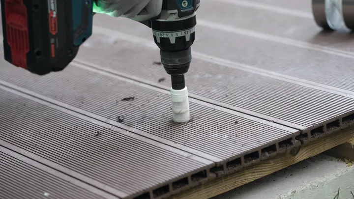 Een persoon die veiligheidsuitrusting draagt, boort met een boormachine een gat in een bruine plank van het terras.