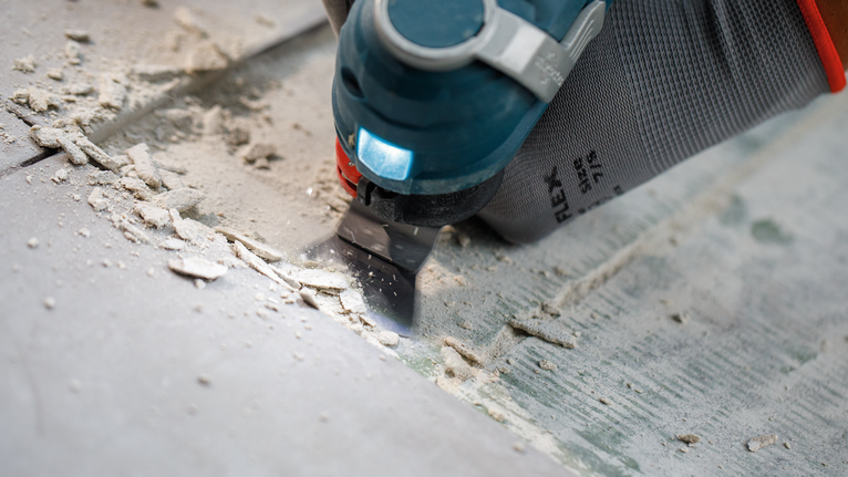 Een persoon met veiligheidsuitrusting verwijdert tegellijm met een multitool.