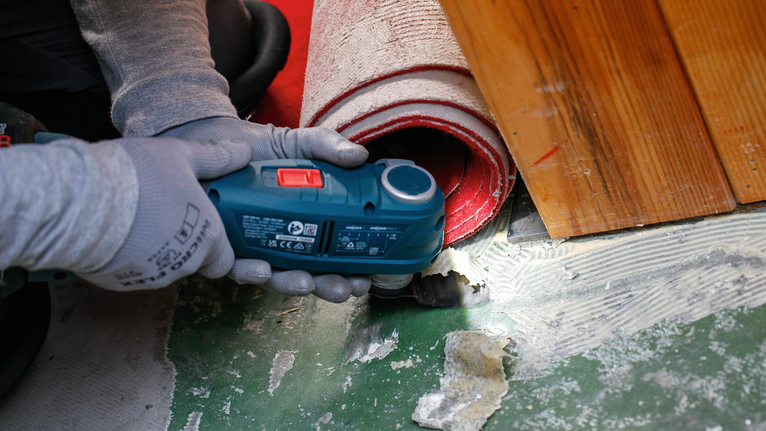 Een persoon die veiligheidsuitrusting draagt, gebruikt een elektrisch gereedschap om de vloerbedekking in de buurt van een opgerold tapijt te snijden.