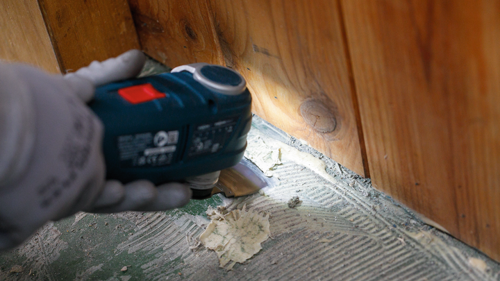 Persoon met veiligheidsuitrusting schraapt oude lijm van de vloer met een elektrisch gereedschap.