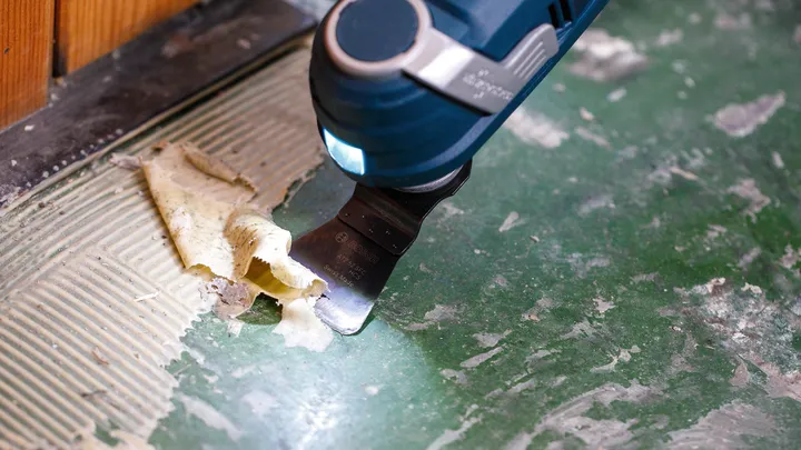 Een elektrisch gereedschap schraapt lijm van een groen vloeroppervlak.