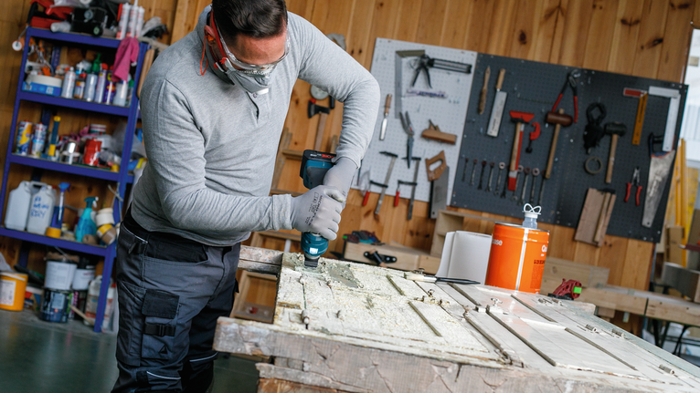 Een persoon met veiligheidsuitrusting boort in een houten deur in een werkplaats.