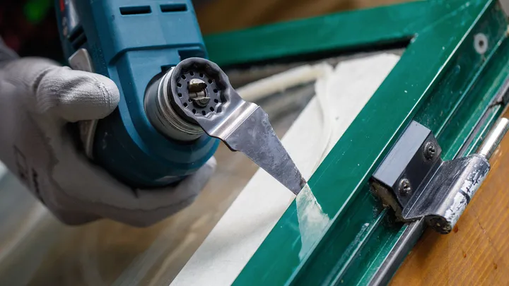 Een persoon met veiligheidsuitrusting verwijdert kit van een raamkozijn met een schraper.