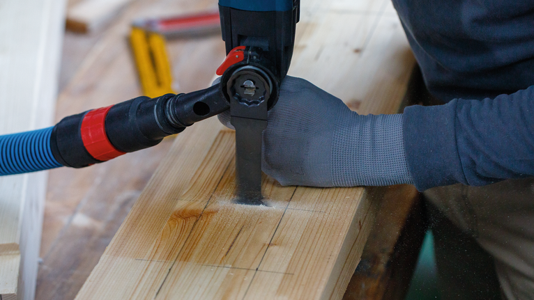 Een persoon die veiligheidsuitrusting draagt, gebruikt een elektrische zaag om een houten plank te zagen.