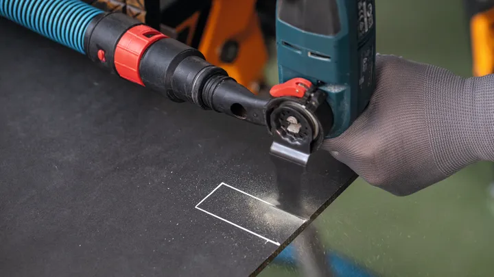 Een persoon met veiligheidsuitrusting snijdt met een elektrisch gereedschap een rechthoekige vorm in een donker bord.