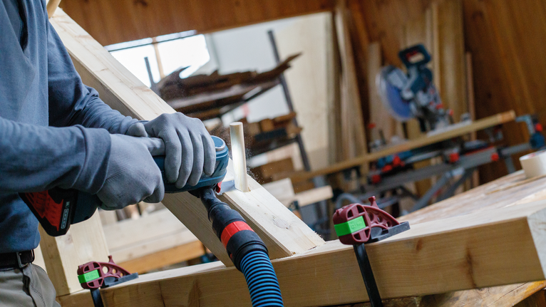 Een persoon met veiligheidsuitrusting boort in een houten plank in een werkplaats.