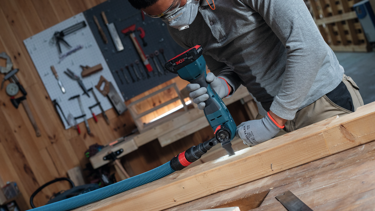 Een persoon met veiligheidsuitrusting boort in een houten balk in een werkplaats.