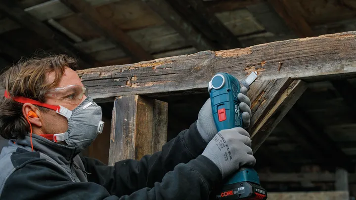 Een persoon die veiligheidsuitrusting draagt, gebruikt een elektrisch gereedschap om een houten balk te schuren.