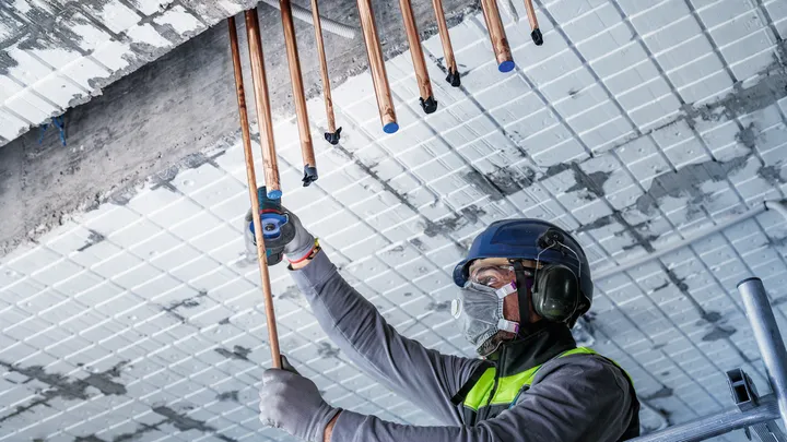 Een persoon met veiligheidsuitrusting installeert koperen leidingen in een plafond.