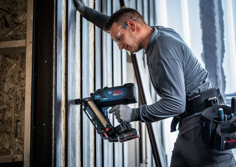 Een persoon die veiligheidsuitrusting draagt, gebruikt een snoerloze tacker op een houten frame.