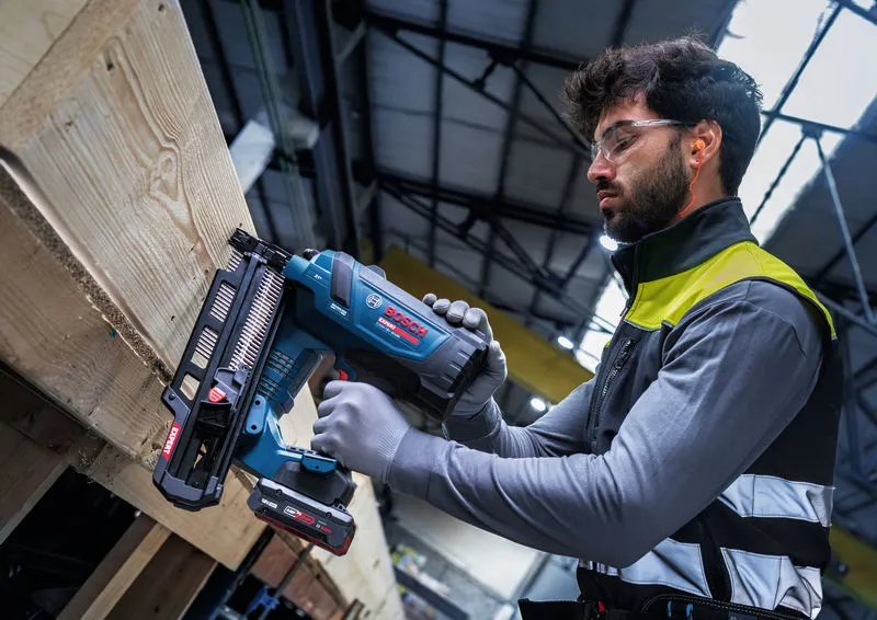 Een persoon die veiligheidsuitrusting draagt, gebruikt een snoerloze houtspijkermachine op een houten balk.