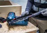 Een persoon met veiligheidsuitrusting laadt spijkers in een snoerloze tacker op een werkbank.