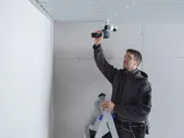 Een persoon die veiligheidsuitrusting draagt, installeert een beveiligingscamera aan het plafond met behulp van een accuboormachine.