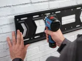 Een persoon bevestigt een metalen beugel aan een witte bakstenen muur met een accuboormachine.
