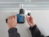 Iemand installeert een beveiligingscamera aan het plafond met behulp van een accuboormachine.