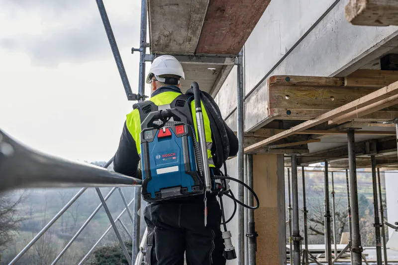 Een persoon met veiligheidsuitrusting staat buiten op een steiger met een blauw rugzakstofzuigersysteem.