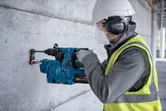 Een arbeider, voorzien van veiligheidsuitrusting, boort met een boorhamer in een betonnen muur.
