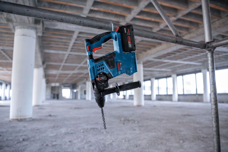 Een draadloze roterende hamerboor hangt aan een metalen steiger in een groot, onafgewerkt gebouw.