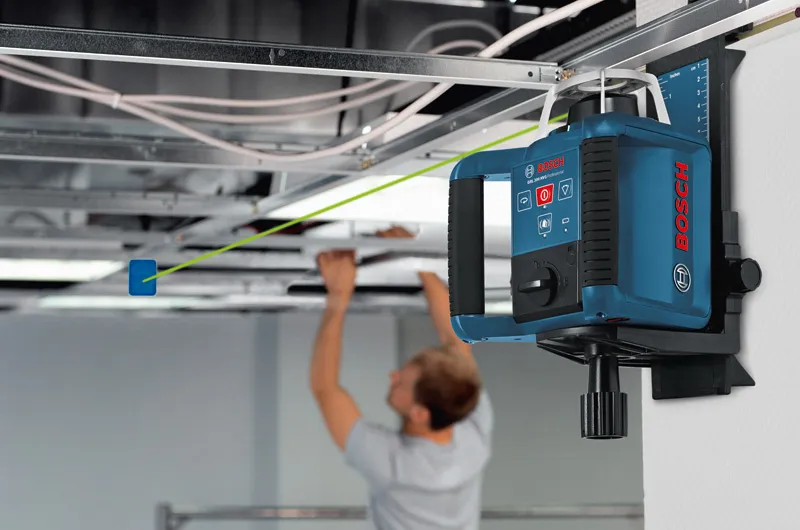 Een laserwaterpas projecteert een groene lijn terwijl iemand een systeemplafond installeert.