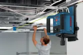 Een laserwaterpas projecteert een groene lijn terwijl iemand een systeemplafond installeert.