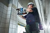 Човек, носещ предпазни средства, пробива в облицована с плочки стена с помощта на безжичен перфоратор.