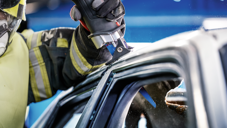 Човек, носещ предпазни средства, използва електрически инструмент, за да реже през рамката на автомобила.