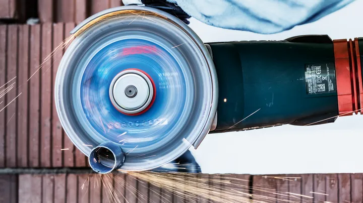 Човек, носещ предпазни средства, реже метална тръба с въртящ се ъглошлайф.