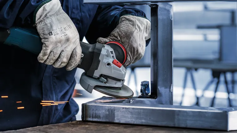 Човек, носещ предпазни средства, шлифова метална греда с ъглошлайф.