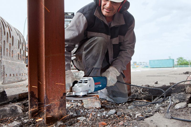 Човек, носещ предпазни средства, използва ъглошлайф, за да реже стоманена греда на открито.