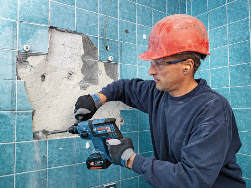 Una persona que usa equipo de seguridad utiliza un martillo rotatorio inalámbrico para quitar azulejos de la pared.