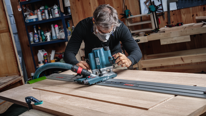 Una persona que usa equipo de seguridad guía una fresadora a lo largo de una pista para dar forma a una tabla de madera.